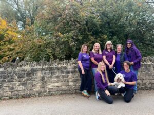 Seven women in purple shirts pose with a small white dog